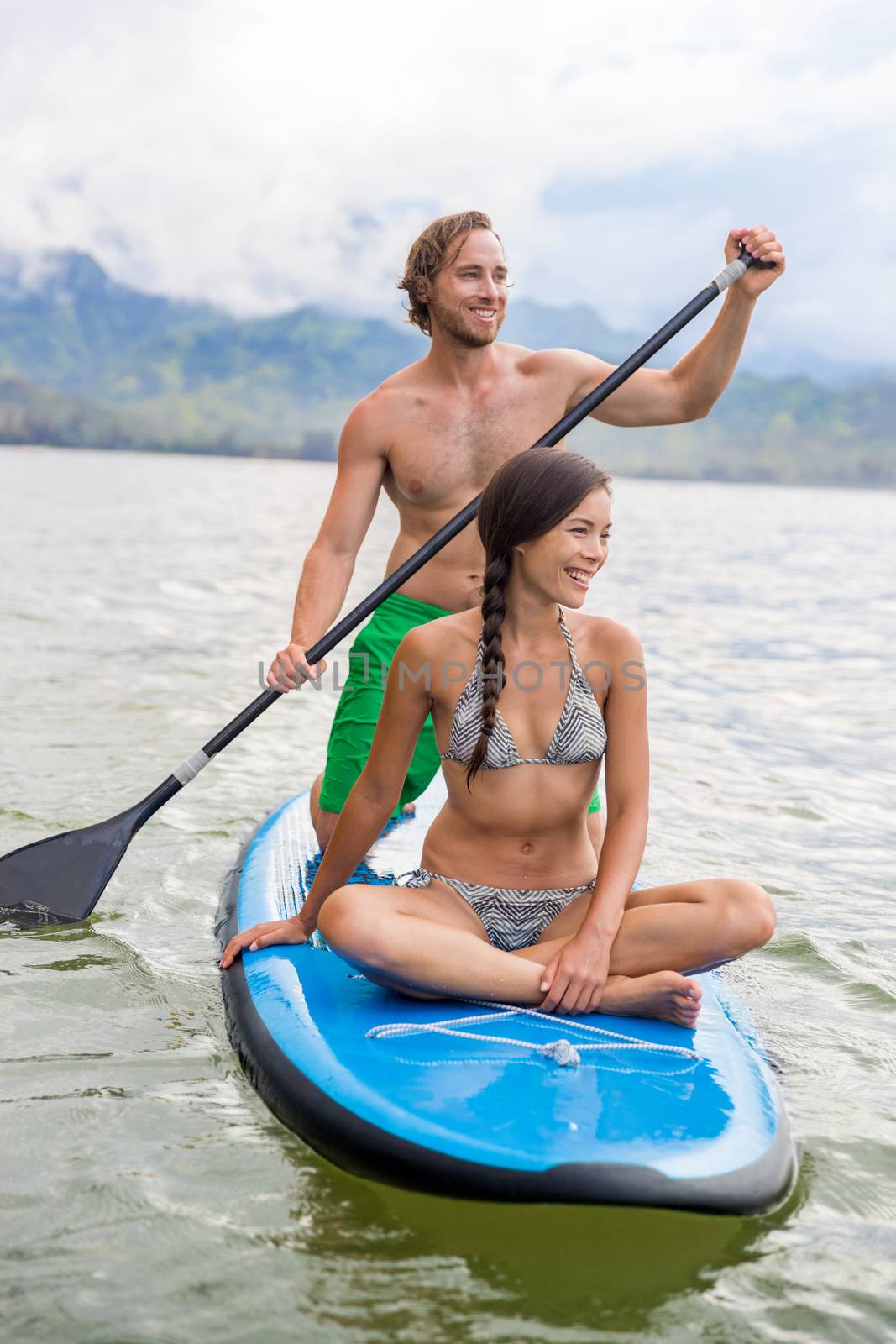 Paddleboard couple having fun paddleboarding together on Hawaii beach vacation summer holidays. Woman sitting on board while man paddles by Maridav