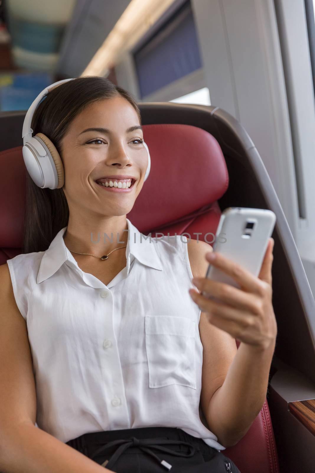 Modern people city lifestyle. Young urban woman using phone app and wireless headphones to listen to music or audiobooks online. Asian girl enjoying train travel in business class seat.