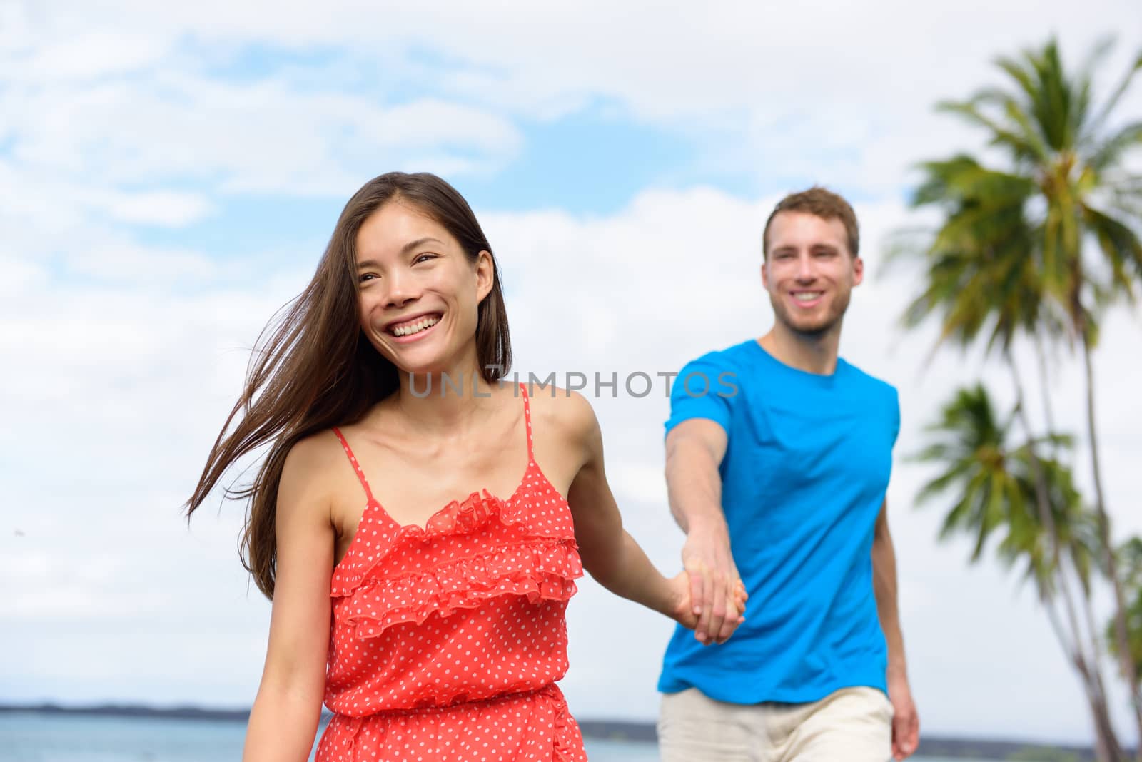 Happy summer vacation couple walking holding hands on beach travel destination. Asian woman caucasian man multiracial people together having fun.