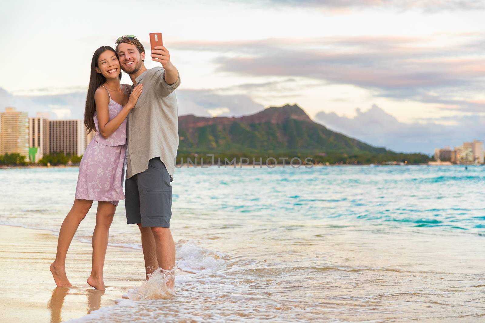 Couple walking on beach at sunset taking selfie picture on mobile phone relaxing together on Waikiki beach, Honolulu, Hawaii travel vacation. Romantic holiday destination for honeymoon by Maridav
