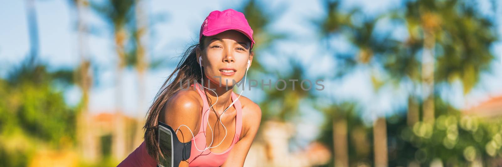 Tired runner woman running with sports cap and sport armband with earphones listening to mobile music. Active fit Asian girl resting taking a jogging break on outdoor summer lifestyle banner panorama.