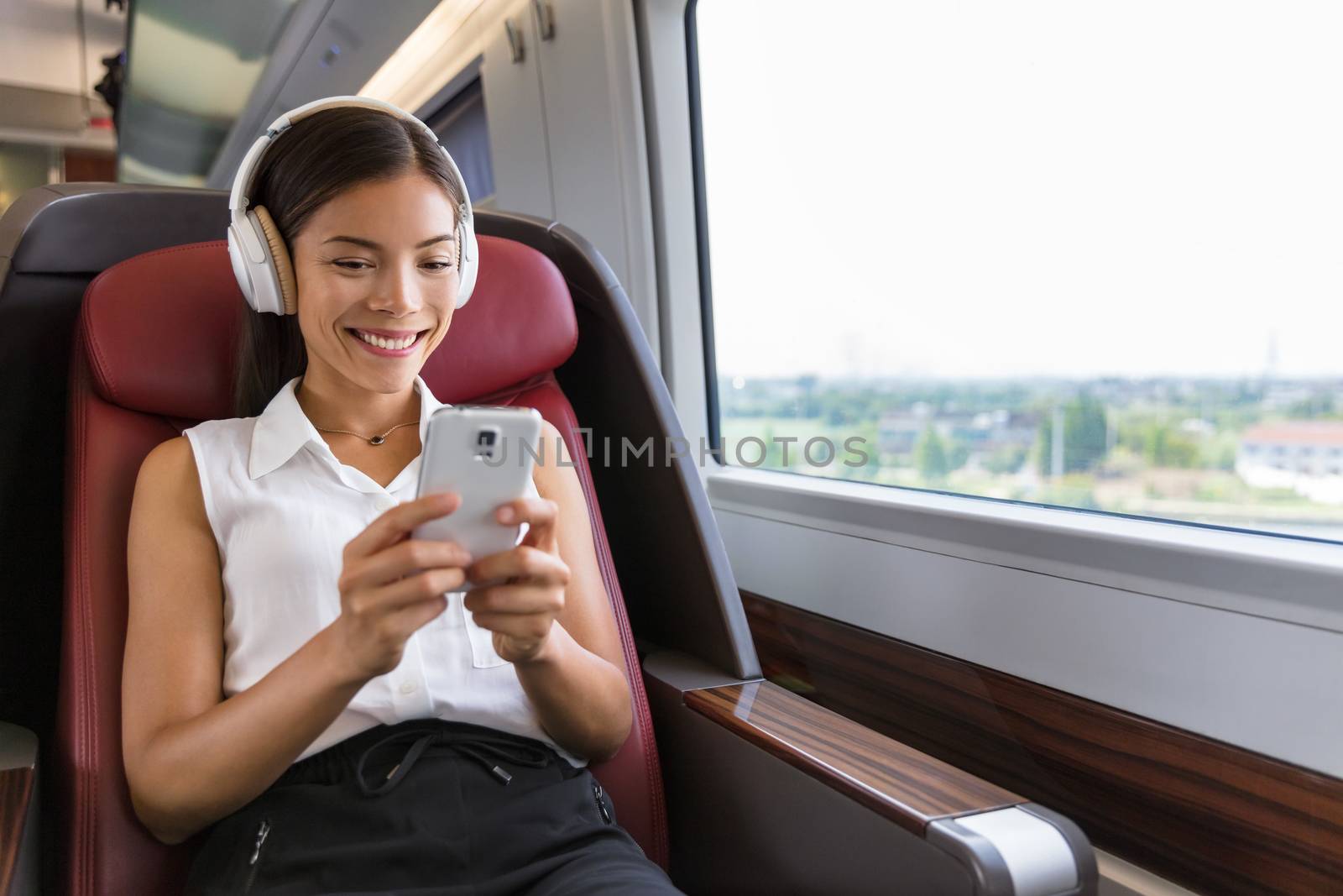 Modern people city lifestyle. Young urban woman using phone app and wireless headphones to listen to music or play video games online. Asian girl enjoying train travel in business class seat.