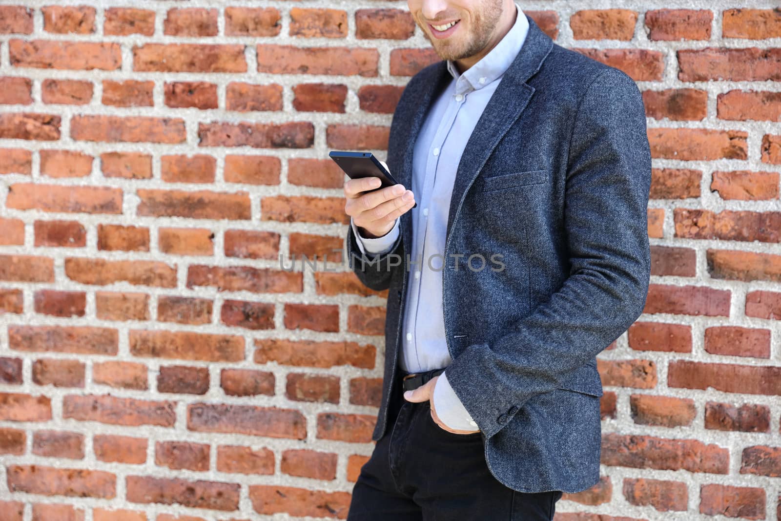 Young casual business man using smartphone in urban city background relaxing on brick wall texting sms on phone app living a modern lifestyle. Closeup of unrecognizable person.