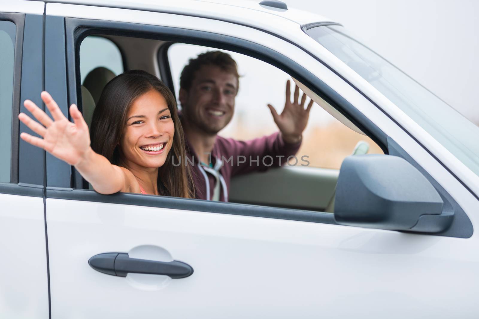 Happy multiracial couple on car travel together on summer vacation. Joyful young people smiling waving hello driving on road trip holidays. New young owners by Maridav