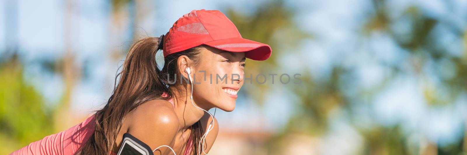 Summer running healthy active woman runner listening to music on phone sport armband with headphones earphones on city street. Happy fitness Asian jogging gIrl with sports cap ready banner panorama by Maridav