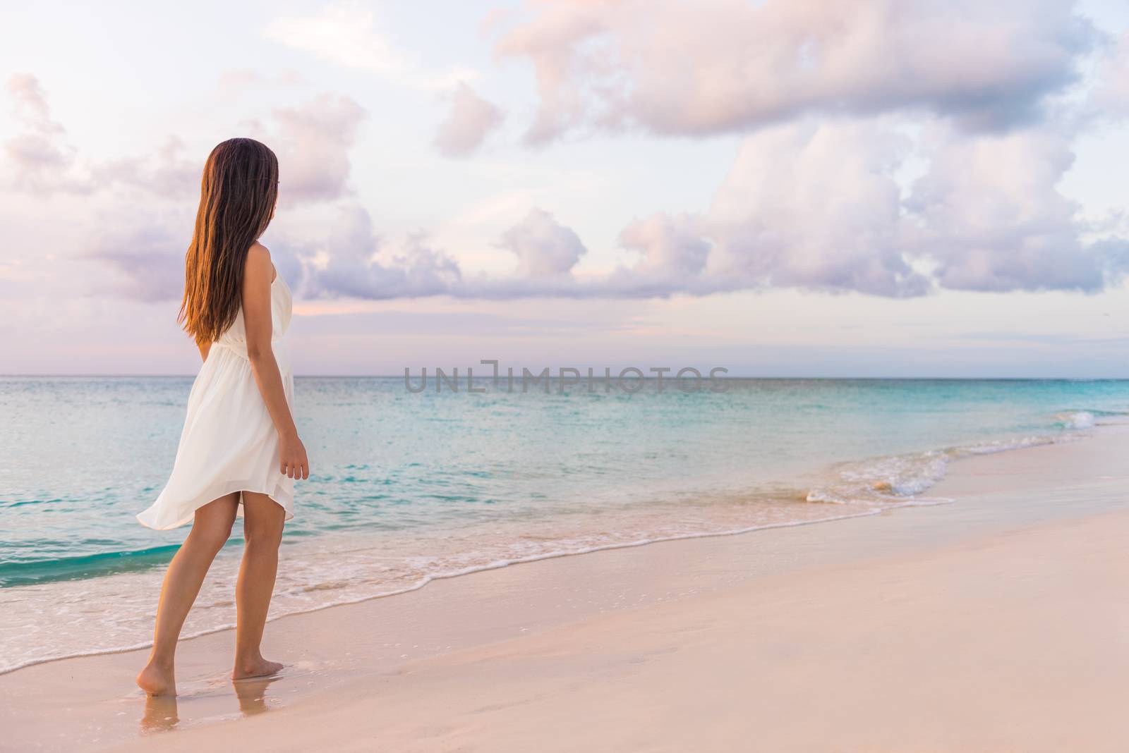 Peaceful vacation paradise woman walking on sunset beach with pastel colors sky and ocean for tranquility and serenity . Girl in white dress relaxing on luxury tropical summer getaway