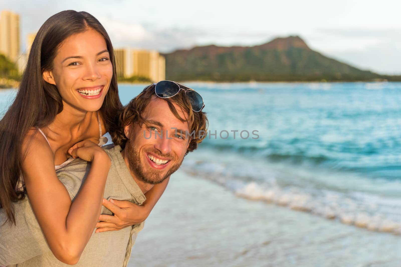 Couple in love having fun piggybacking on Hawaii beach. Healthy people portrait girlfriend and boyfriend hugging happy at sunset on Waikiki vacation, Honolulu, Hawaii. Multiracial relationship by Maridav
