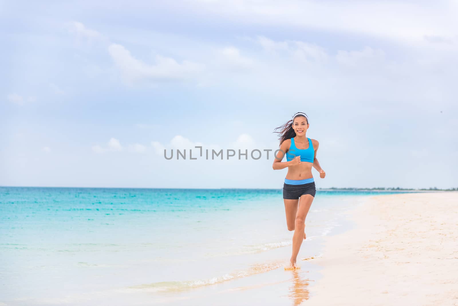 Exercise fitness athlete woman jogging on beach training cardio in summer vacation background. Blue ocean water, sun sky landscape. Happy Asian girl running barefoot outside.