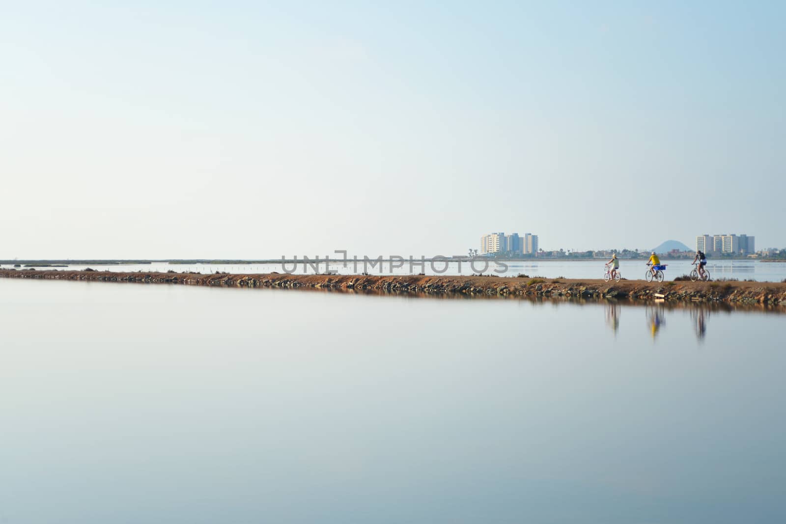 Gentle evening colors surround 3 pedal bike cyclists as they cycle along a thin strip of land between lagoons. Reflections shimmer in still water.
