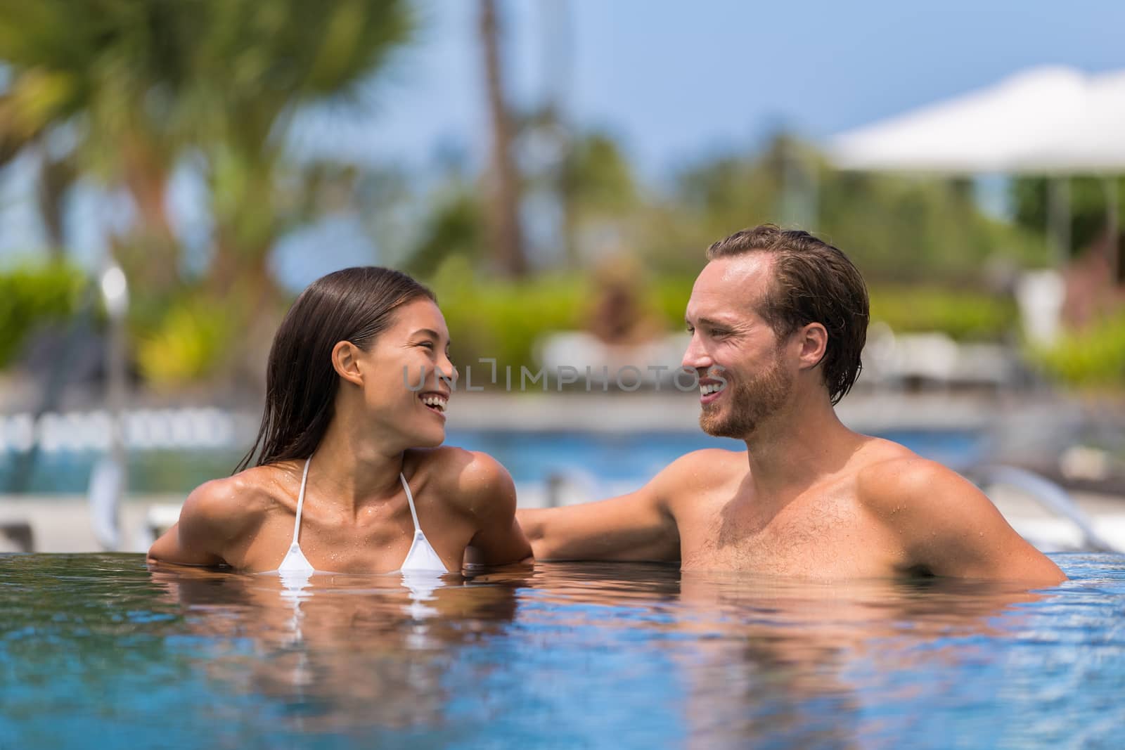 Happy swimming pool couple having fun laughing talking on luxury resort hotel travel holiday together on summer vacation. Asian woman, caucasian man in love on honeymoon by Maridav