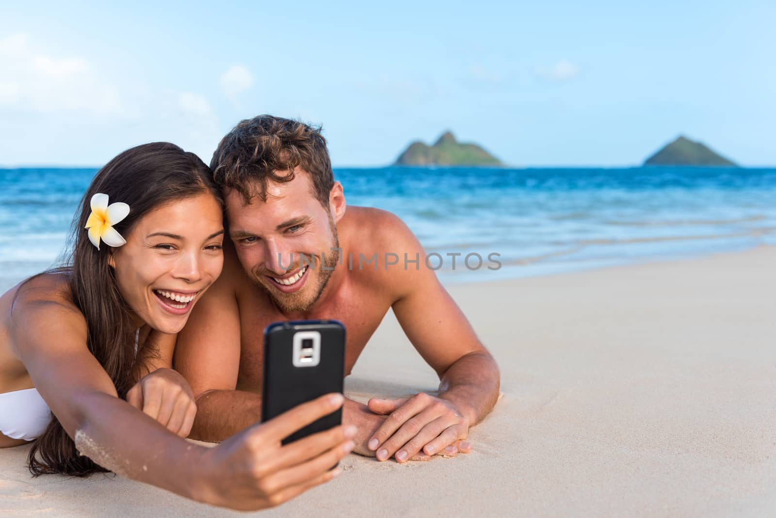 Beach vacation couple taking fun phone selfie on Hawaii vacation. Asian girl Caucasian man relaxing on Lanikai beach, Oahu, on summer holidays using smartphone for pictures together by Maridav