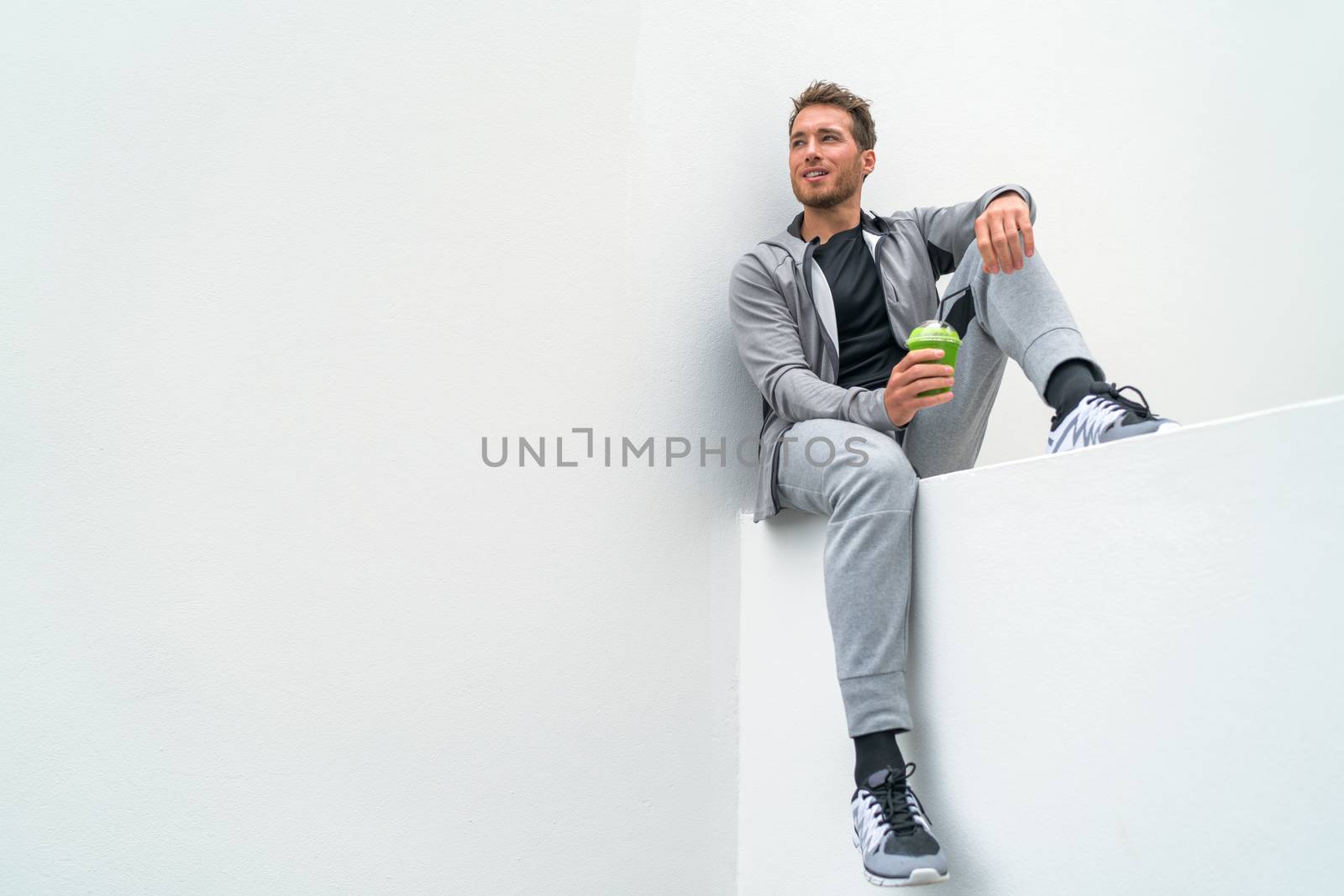 Healthy sport man drinking green smoothie during training at gym in activewear sweatpants outdoor. Athlete sitting relaxing post-workout.