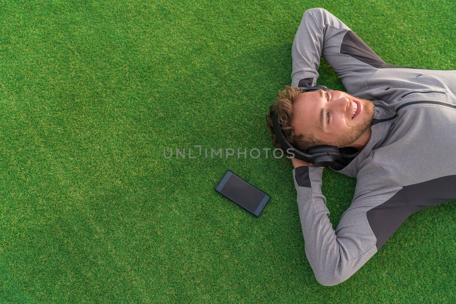 Happy man listening to music with headphones and phone relaxing on green grass lying down with arms behind head enjoying summer day in park. Young adult using mindfulness smartphone app concept.