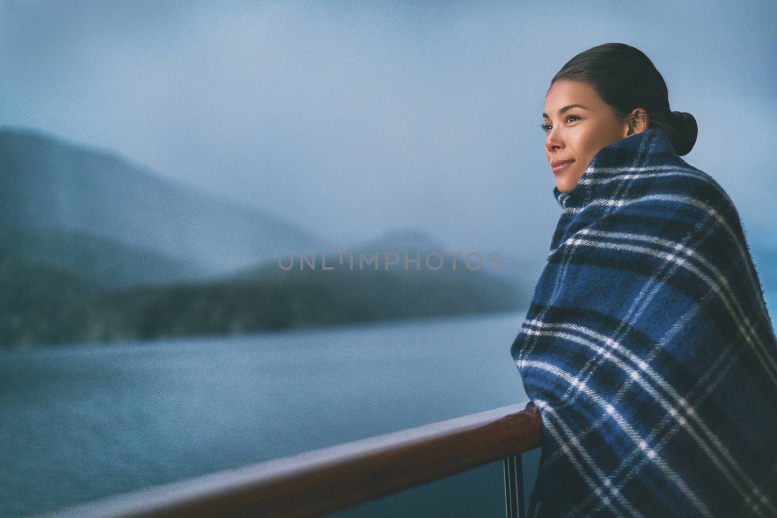 Cruise ship passenger on Alaska travel vacation enjoying scenery at dusk on suite balcony deck with wool throw in cold weather. Asian tourist woman relaxing on summer holiday cruising adventure.