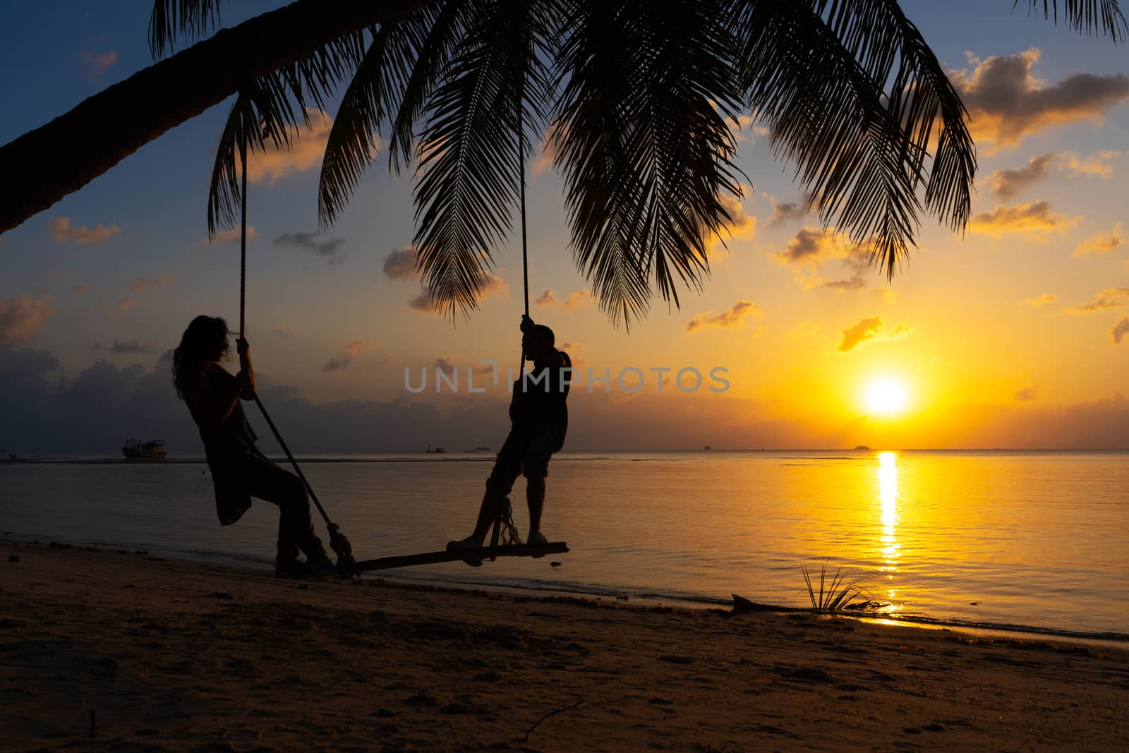 Silhouetted couple in love walks on the beach during sunset. Riding on a swing tied to a palm tree and watching the sun go down into the ocean by Try_my_best