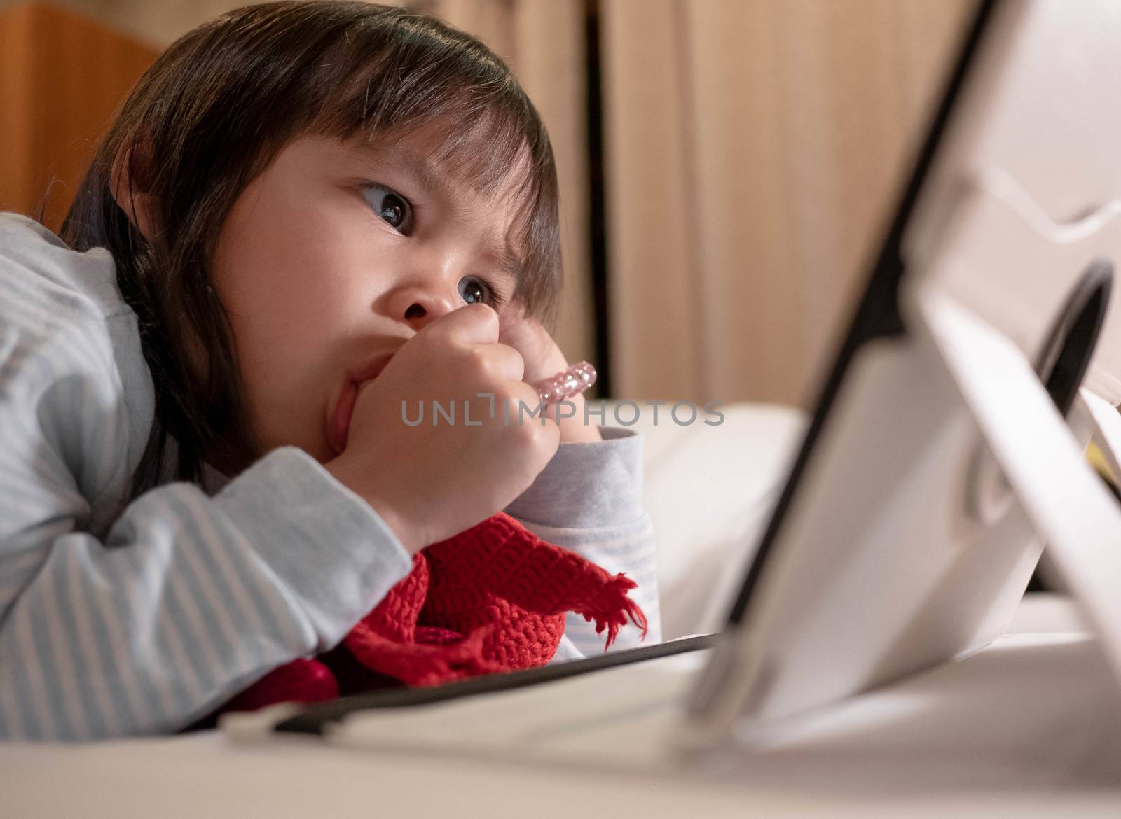 Cute little girl lying on bed with sucking her thumb and looking on a digital tablet in bedroom.