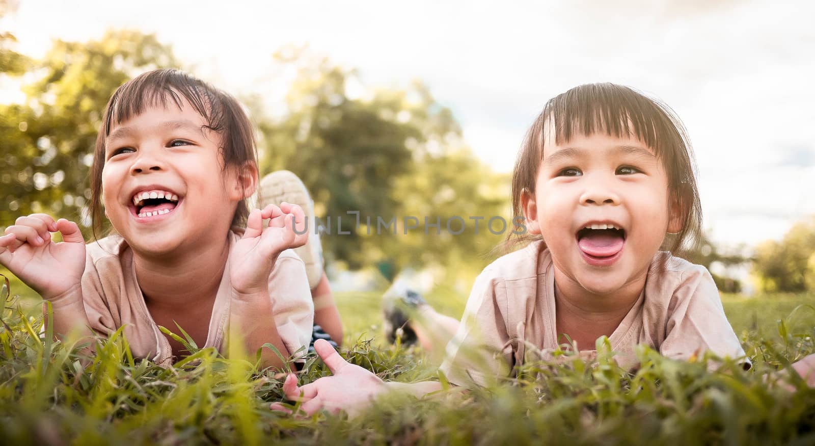 Two happy sibling girls lying on green grass outdoors in summer park.