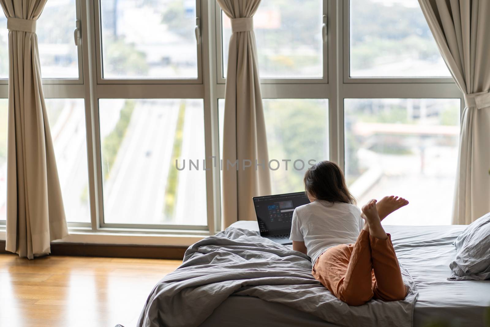 A beautiful young brunette girl is working on the laptop while lying on a bed by a panoramic window with a beautiful view from a high floor. Stylish modern interior.Shopping on the Internet.