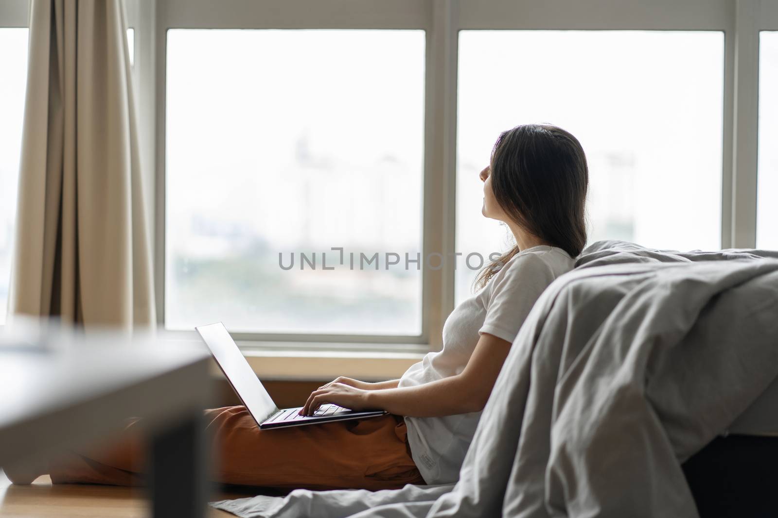 Beautiful young brunette girl working on a laptop, sitting on the floor near the bed by the panoramic window. Stylish modern interior. A cozy workplace. Shopping on the Internet. by Try_my_best