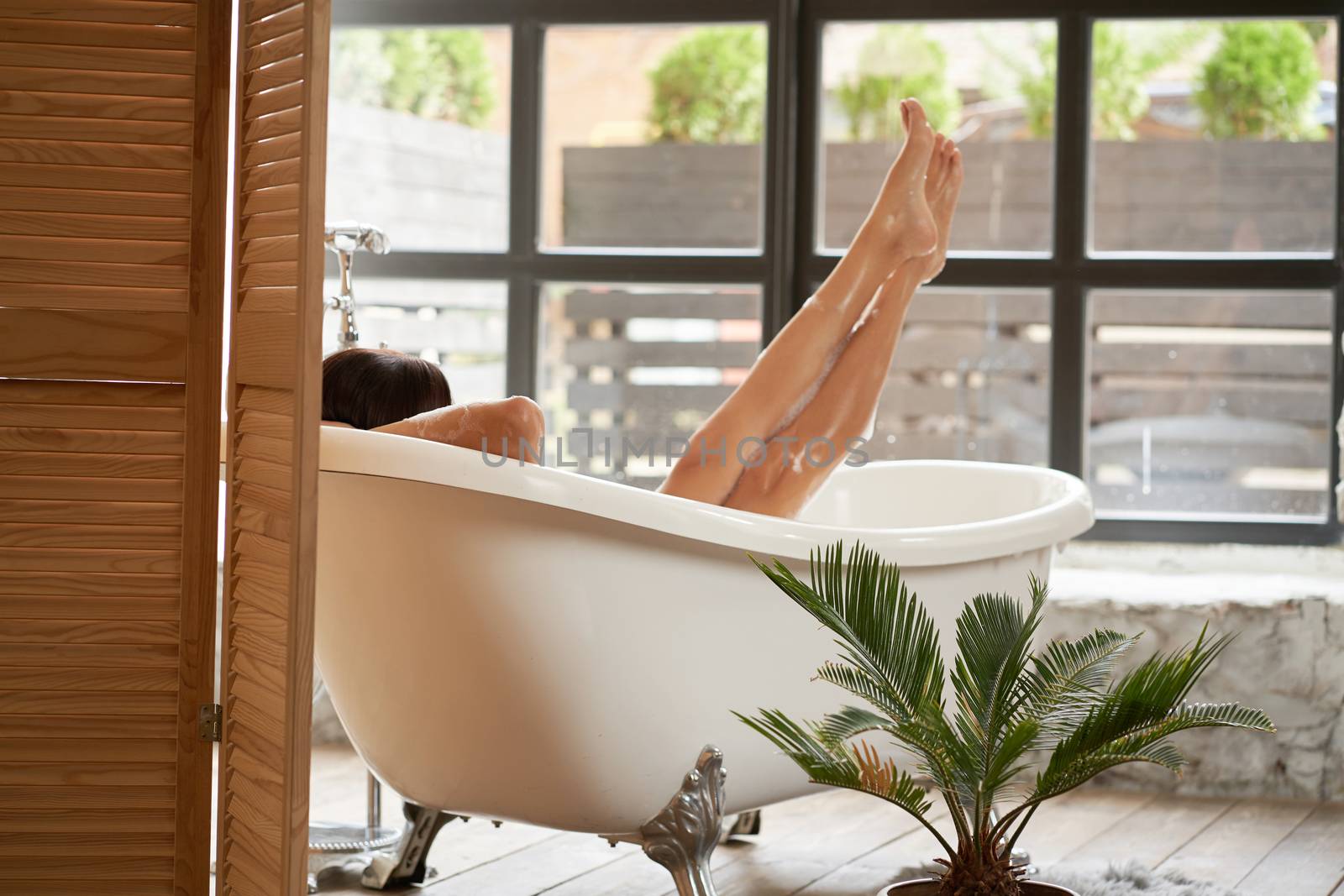 A gorgeous woman is enjoying herself in a white bathtub, in a bright room with a large window