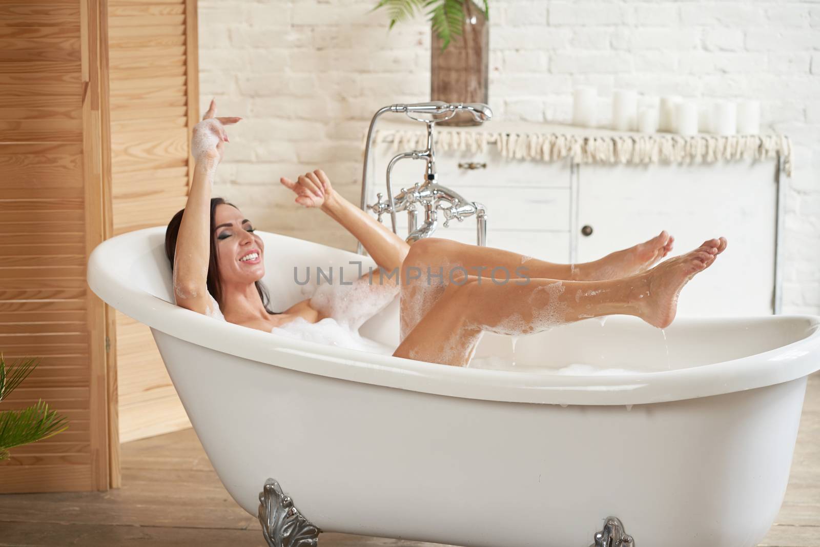 A gorgeous woman is enjoying herself in a white bathtub, in a bright room with a large window