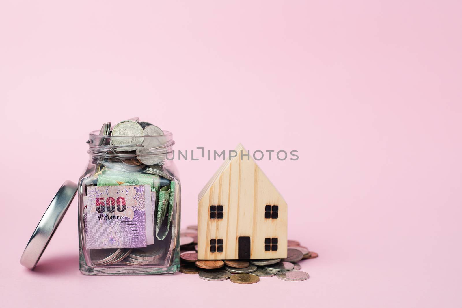 Wooden house model with Thai currency banknote and money coins in the glass jar on pink background for business, finance and property investment concept