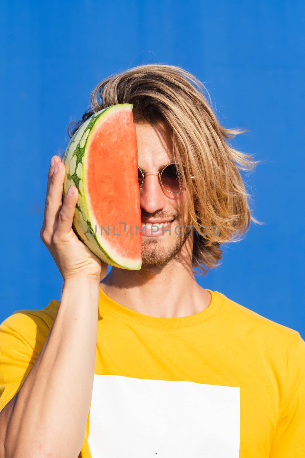 Attractive young man with long blond hair with half a watermelon covering his face on a plain blue background. Summer, sun, heat, fruit, vacation concept.