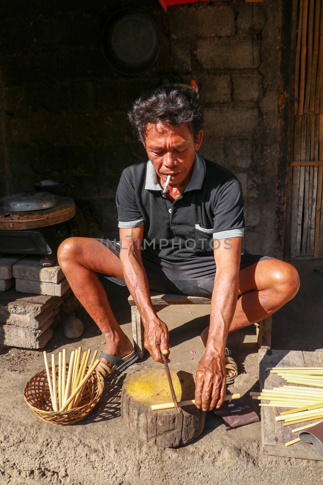 Old man is cutting the bamboo in his workshop to make bamboo straws, Bali, Indonesia. Front view to a worker who makes bamboo straws.