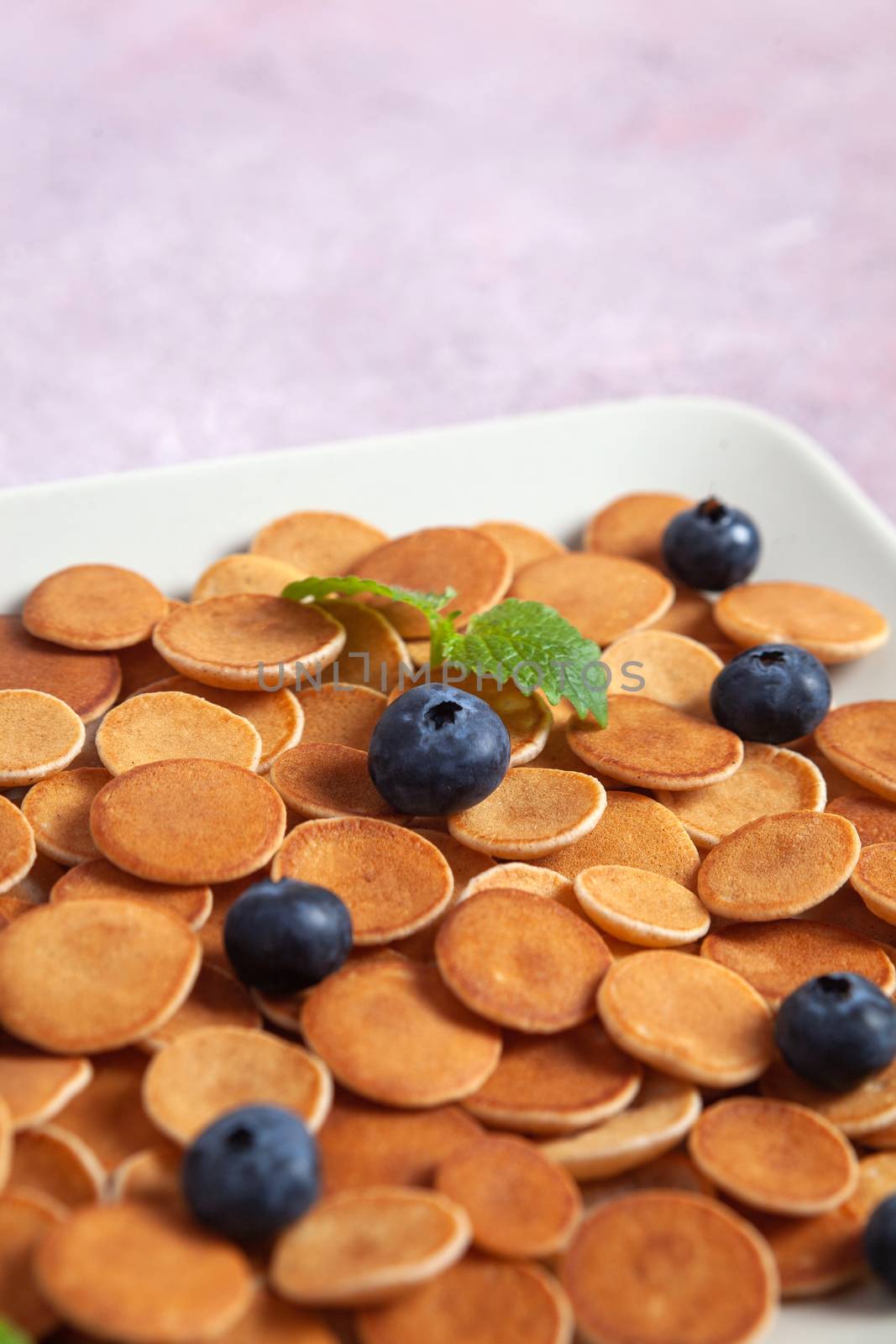 Trendy homemade breakfast, pancake cereal, mini pancakes on light plate on pink concrete background with blueberry, leaves of lemon balm and peppermint. Space for text