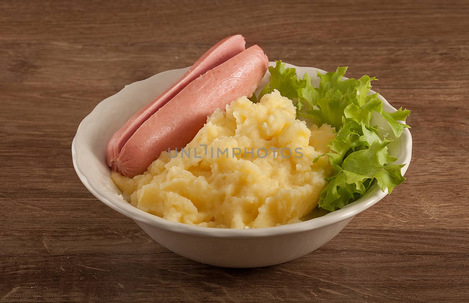 White bowl with mashed potatoes, boiled small sausage and fresh green lettuce on the wooden table