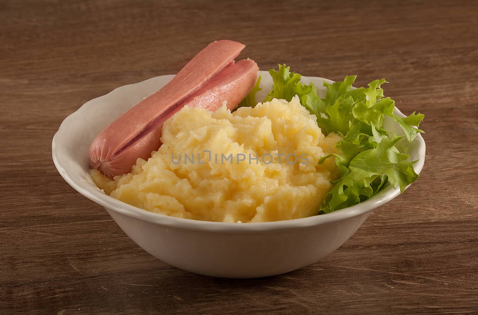 White bowl with mashed potatoes, boiled small sausage and fresh green lettuce on the wooden table