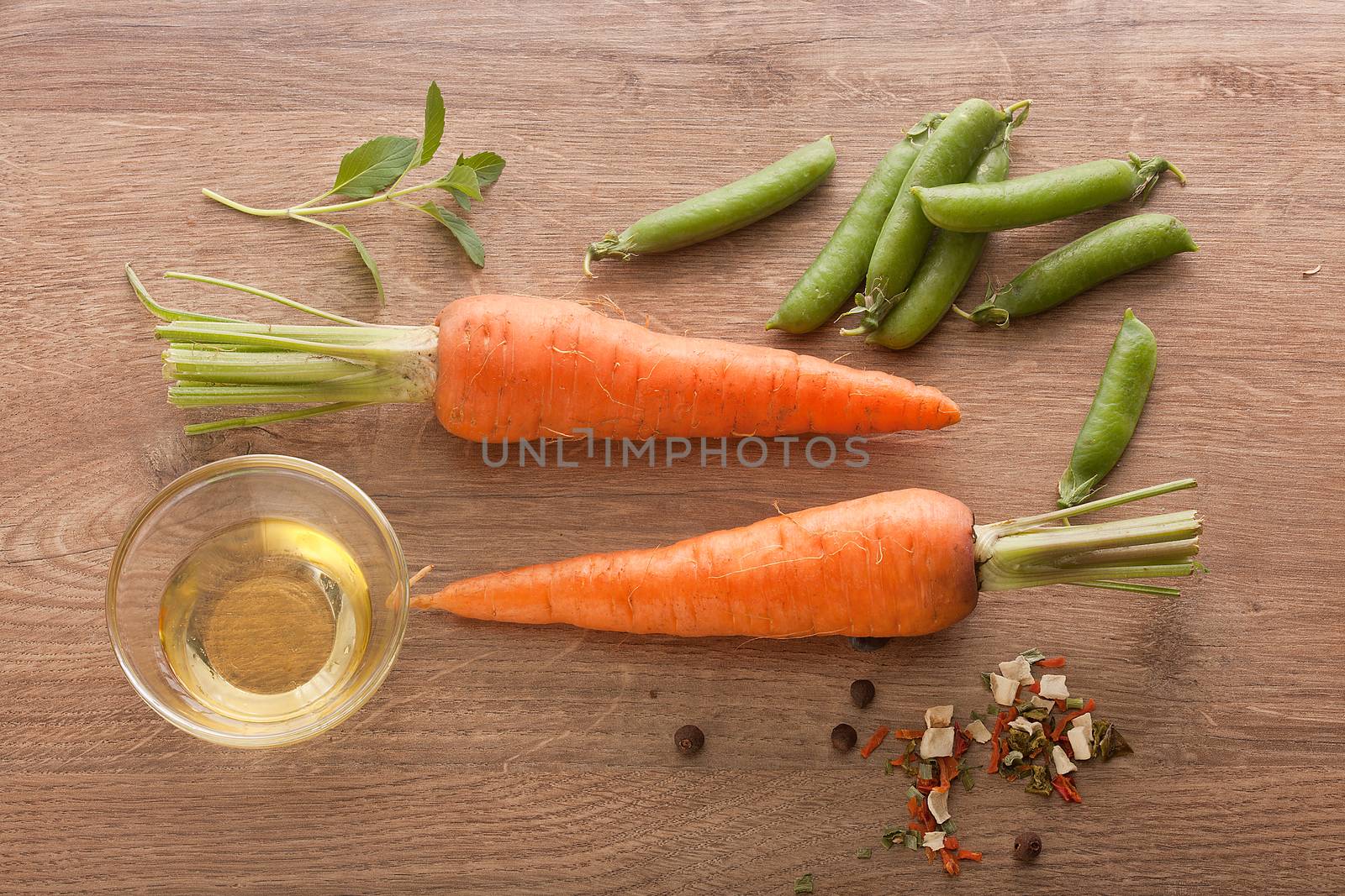 Top view of two whole carrots, fresh green pea pods, oil and dry herbs on the wooden board