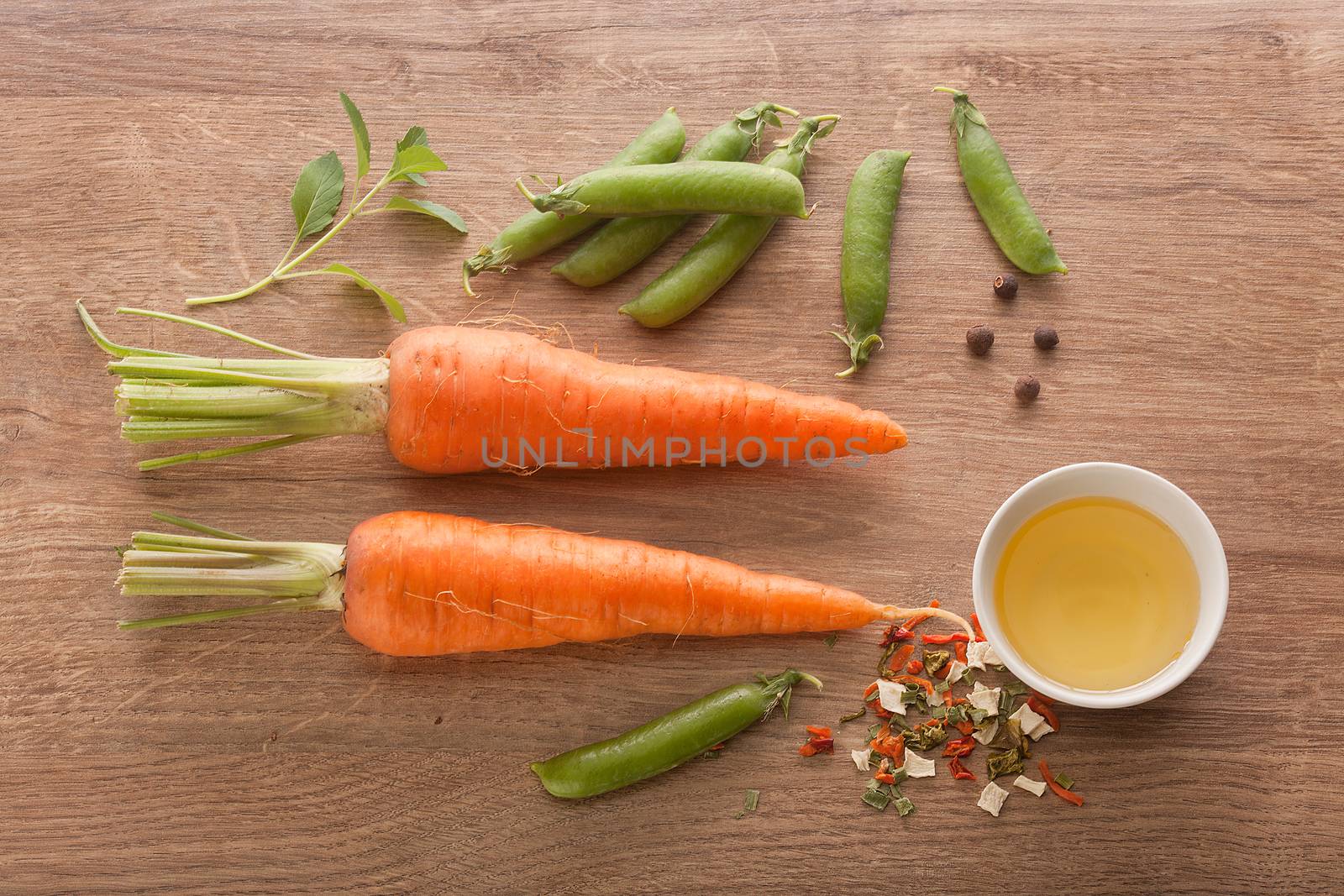 Top view of two whole carrots, fresh green pea pods, oil and dry herbs on the wooden board