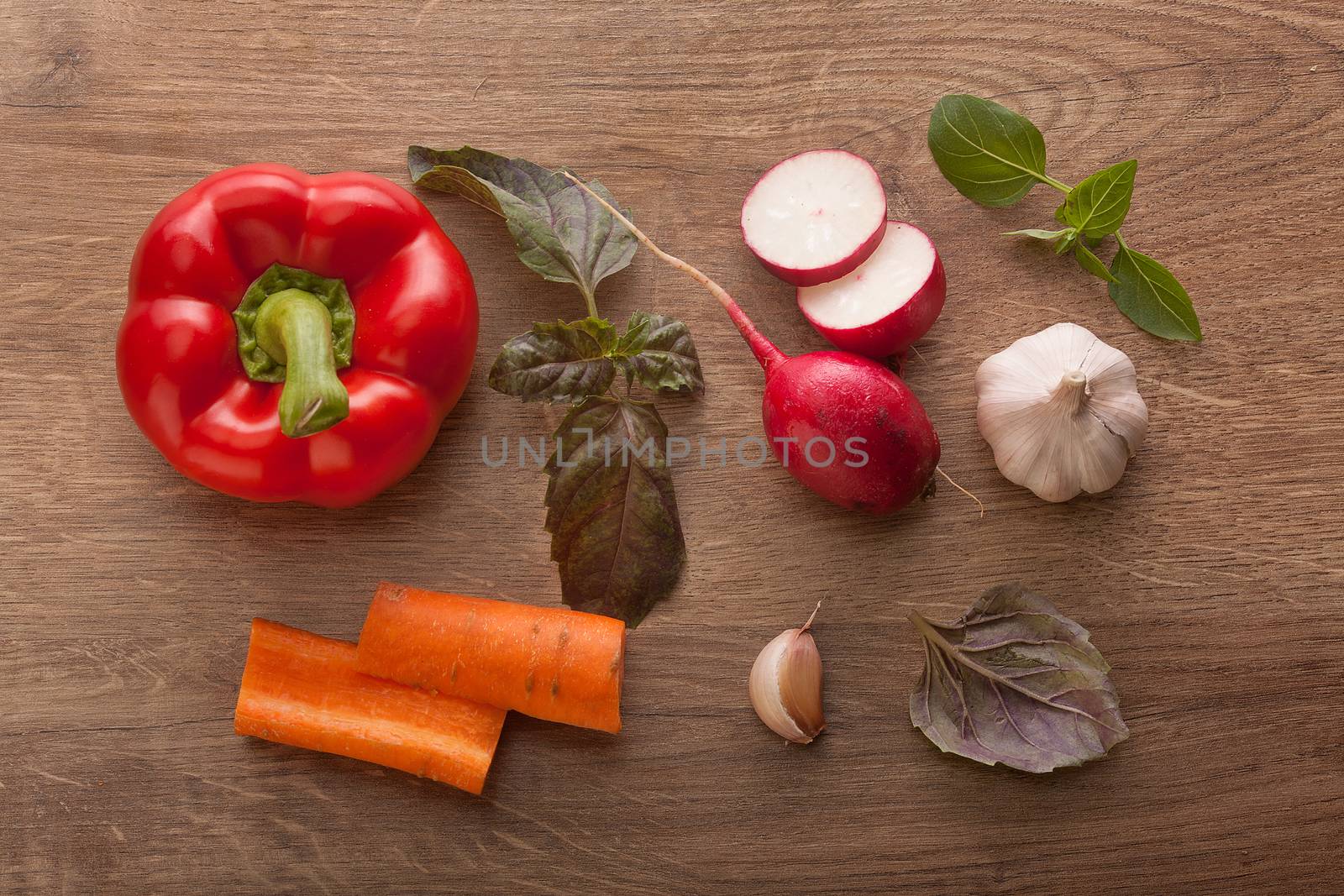 Top view of red paprika, sliced radish, two pieces of carrot, garlic and leaves of basil on the wooden table