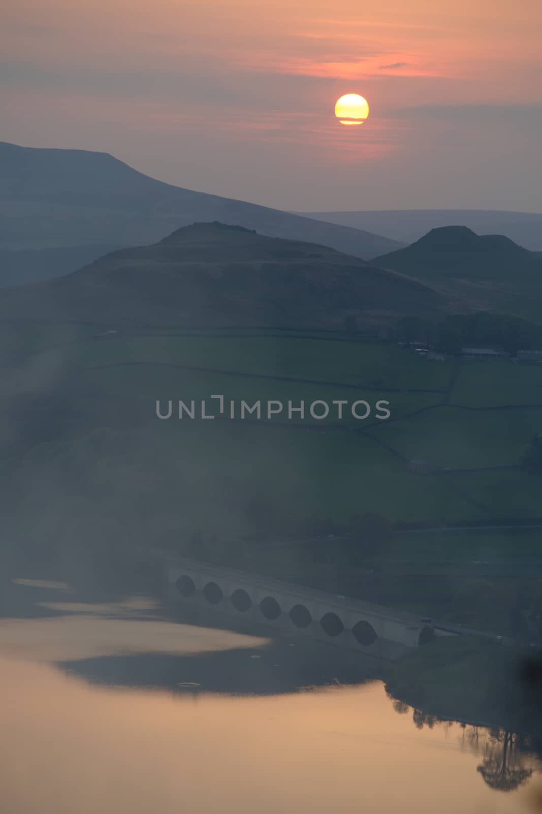 Calm Ladybower reservoir water beneath an arched bridge reflects the orange sunset from above the tall hills.
