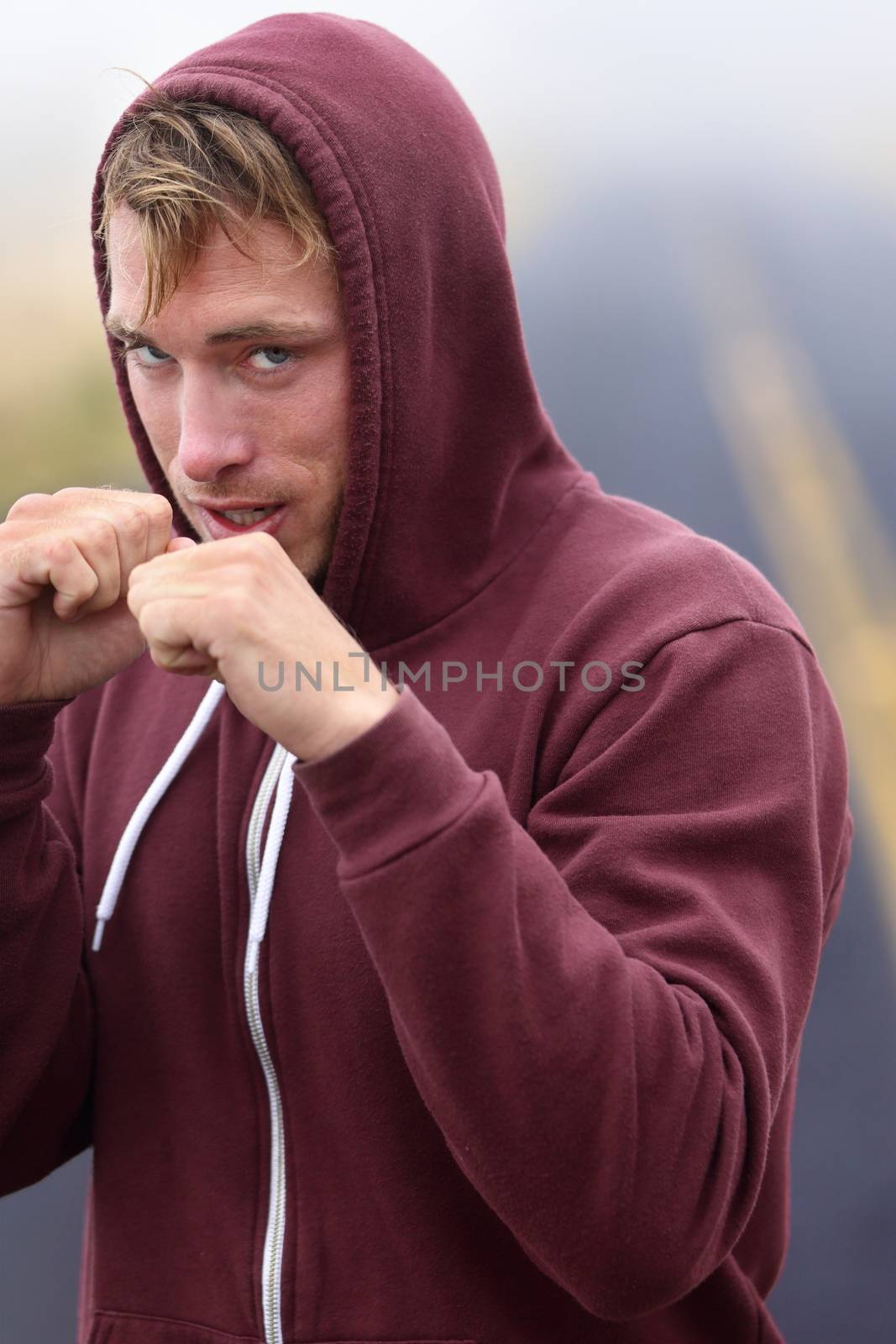 Fitness boxer boxing outside on road in hoodie. Man punching and hitting looking at camera. Fit fitness boxer sweating in sweatshirt outside. Caucasian male fitness model in his 20s.