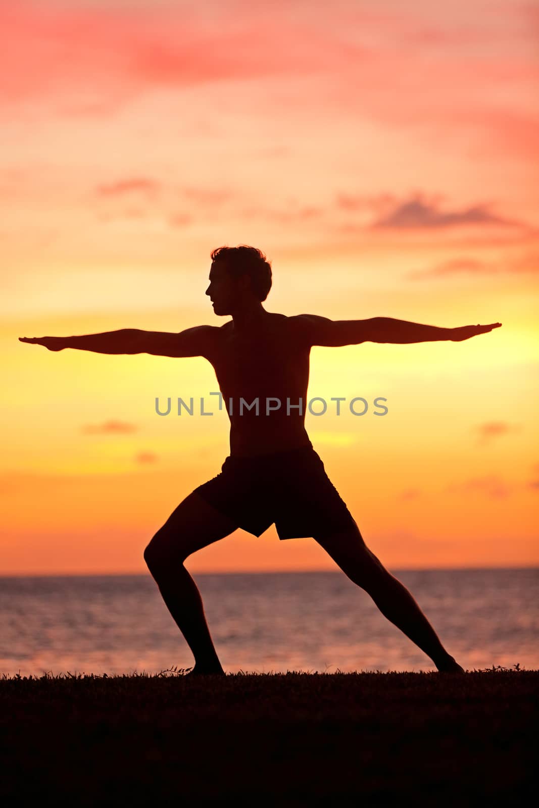 Yoga man training and meditating in warrior pose outside by beach at sunrise or sunset. Male yoga instructor working out training in serene ocean landscape. Silhouette of man model against sun.