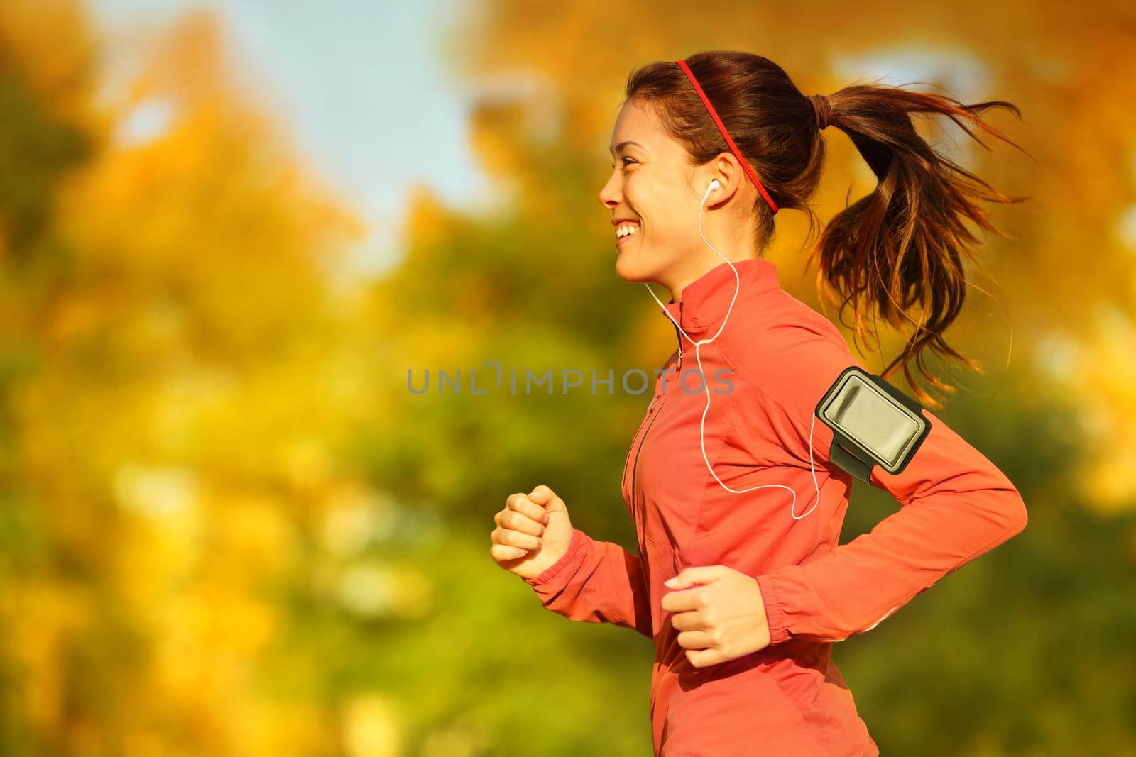 Woman runner running in fall autumn forest listening to music on smartphone using earphones. Female fitness girl jogging on path in amazing fall foliage landscape nature outside.