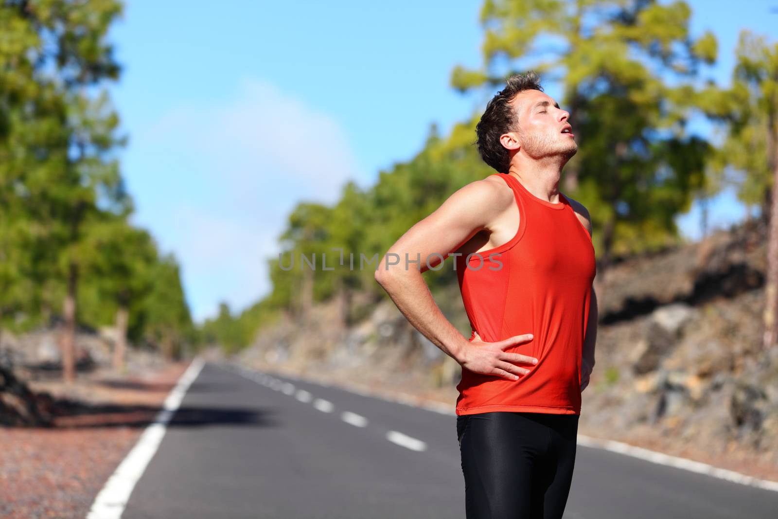 Runner resting tired and exhausted after running. Jogging man taking a break during training outdoors in on mountain road. Young Caucasian male fitness model after work out.