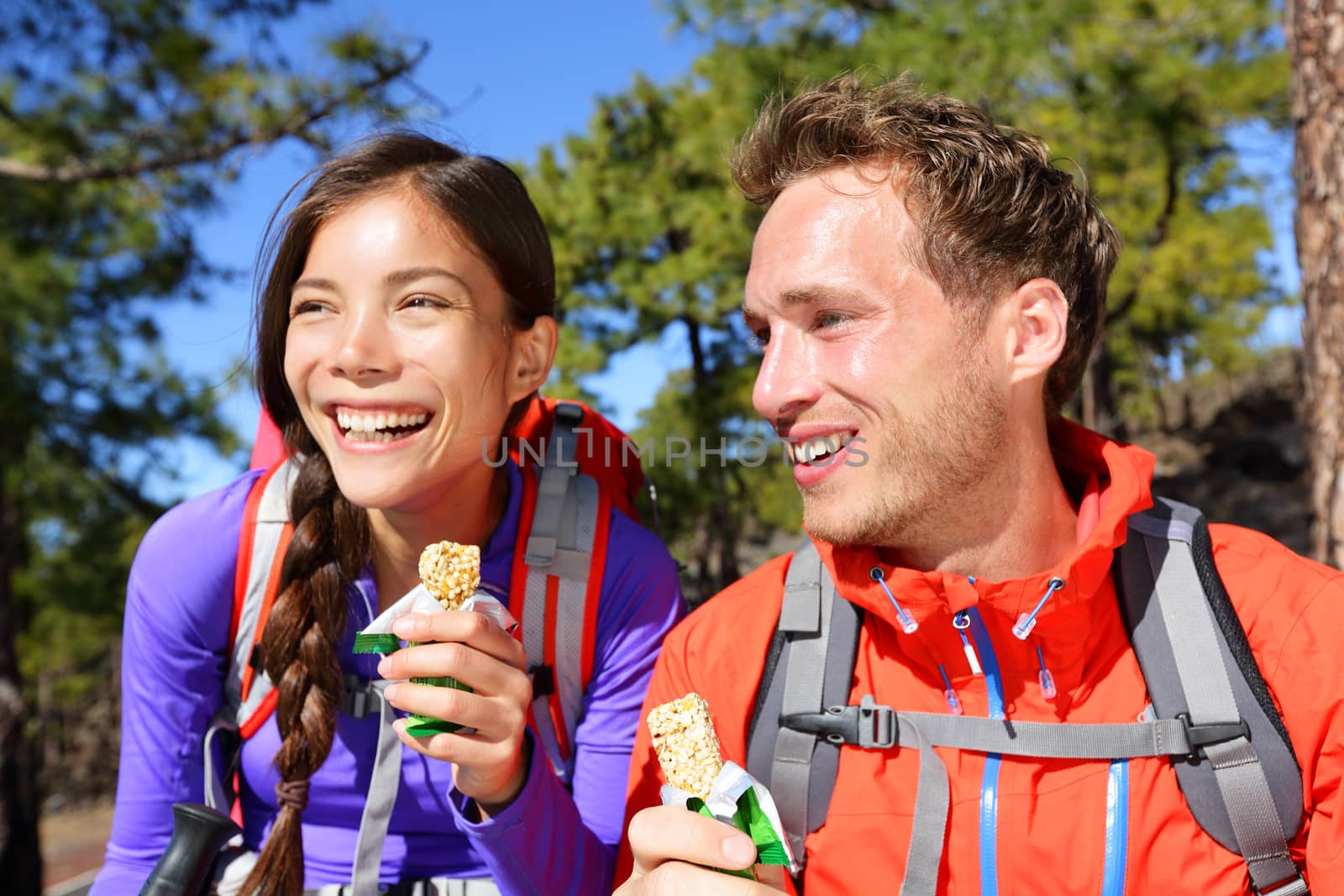 Couple eating muesli bar hiking. Happy people enjoying granola cereal bars living healthy active lifestyle in mountain nature. Woman and man hiker sitting laughing during hike.