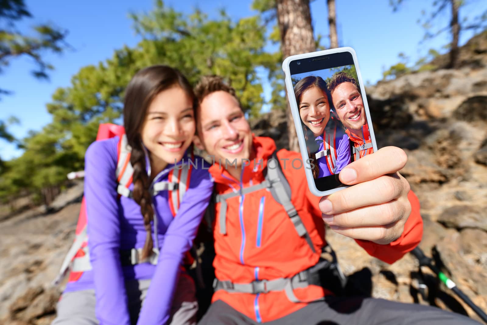 Selfie couple using smart phone hiking in nature with smartphone. Happy couple taking self-portrait photo picture using app. Man and woman having fun together. Focus on screen.