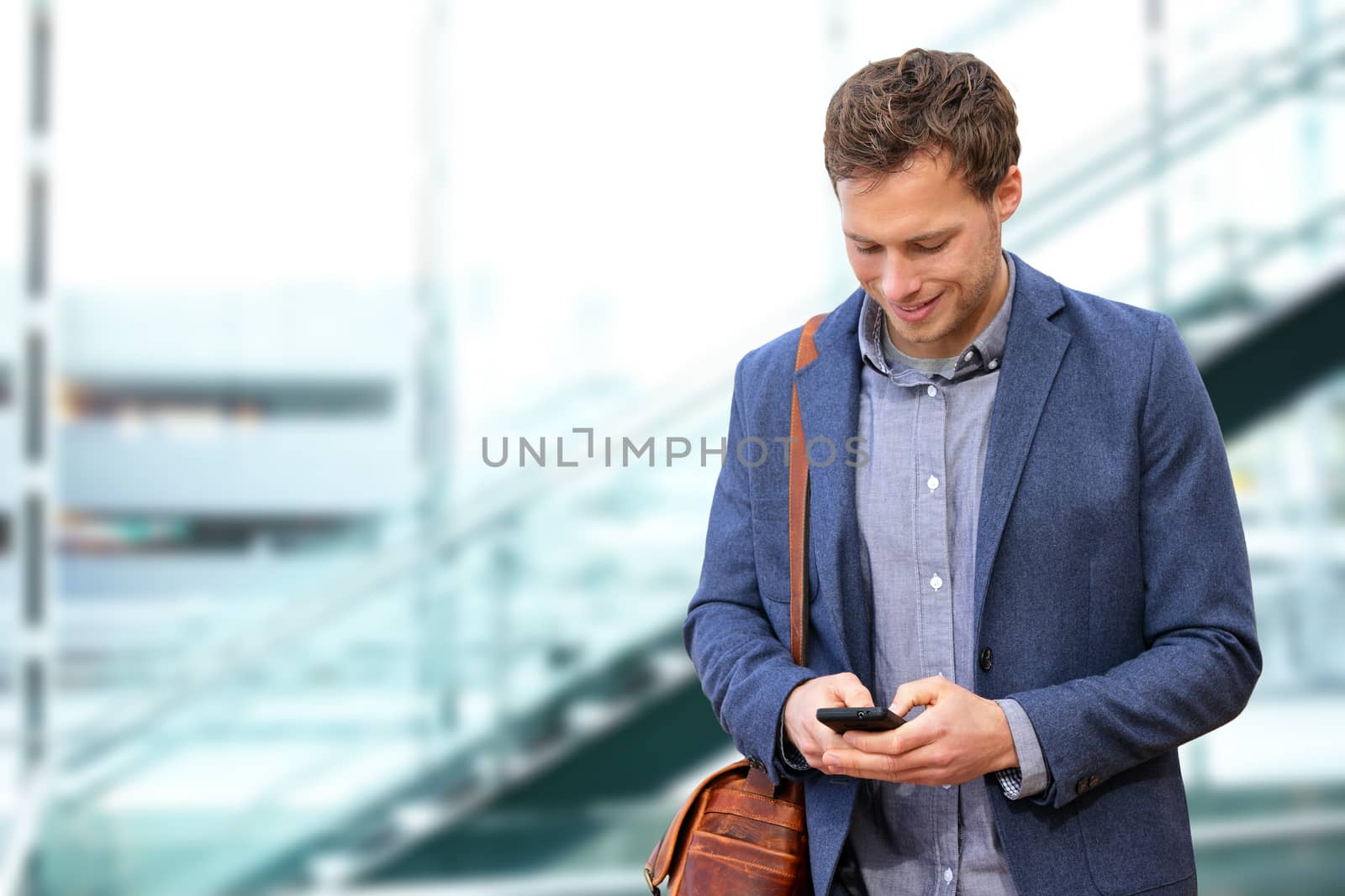 Young urban professional man using smart phone in office building indoors. Businessman holding mobile smartphone using app texting sms message wearing suit jacket and bag.