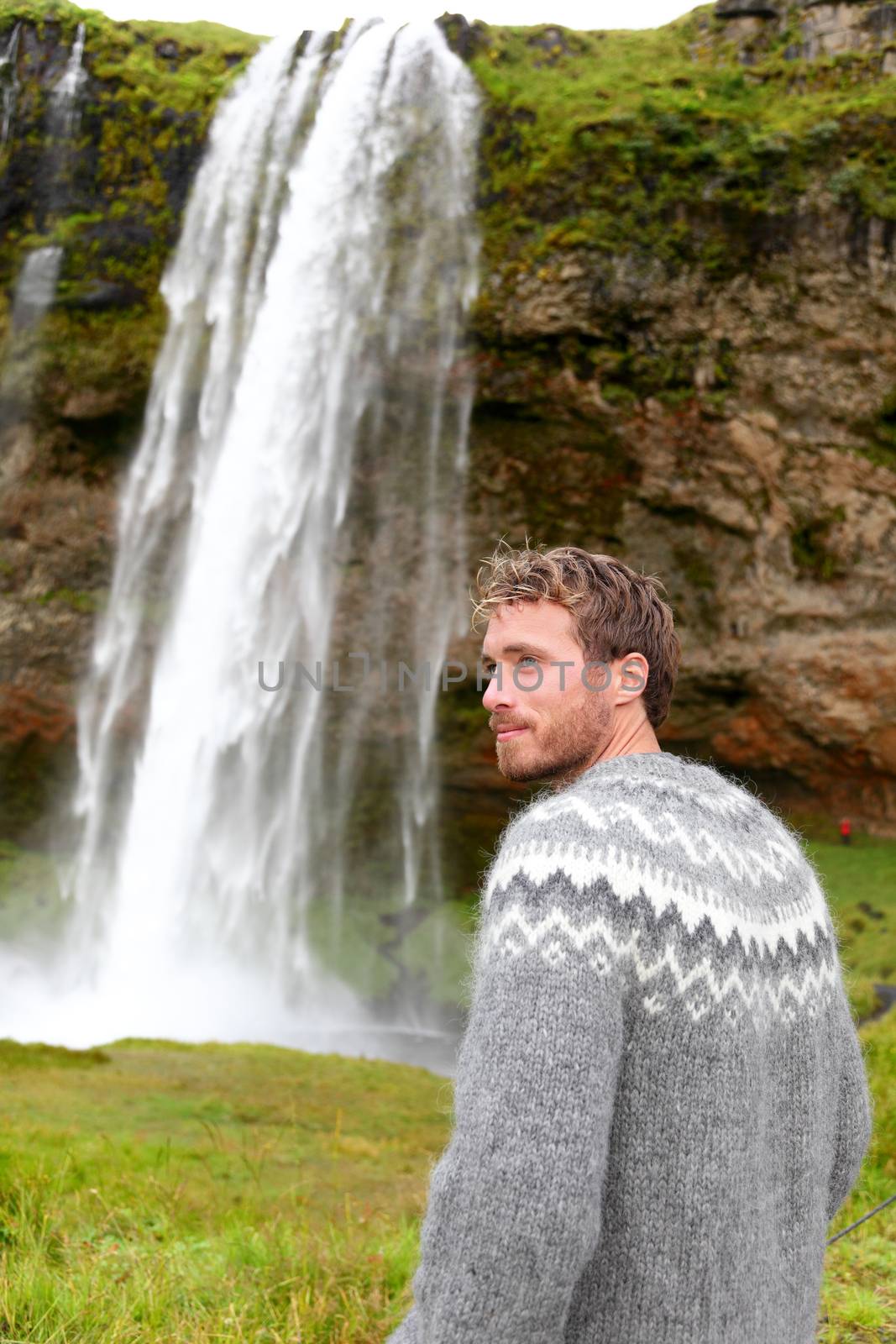 Man in Icelandic sweater by waterfall on Iceland outdoor smiling. Portrait of good looking male model looking to side in nature landscape with tourist attraction Seljalandsfoss waterfall on Ring Road.