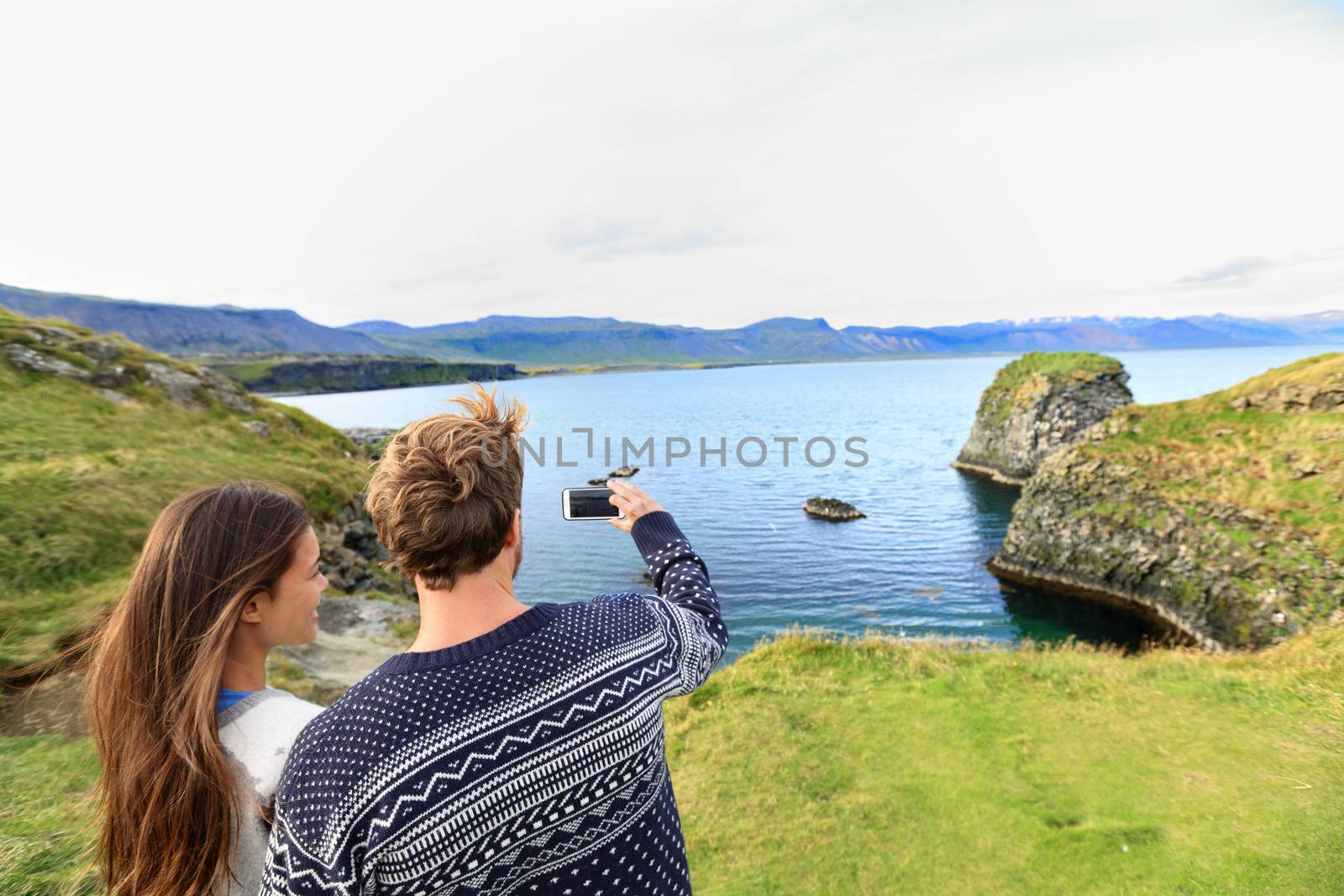 Tourists on travel taking photo with smartphone on Iceland. Happy couple sightseeing taking pictures using smart phone visiting Arnarstapi, Snaefellsnes, Iceland.