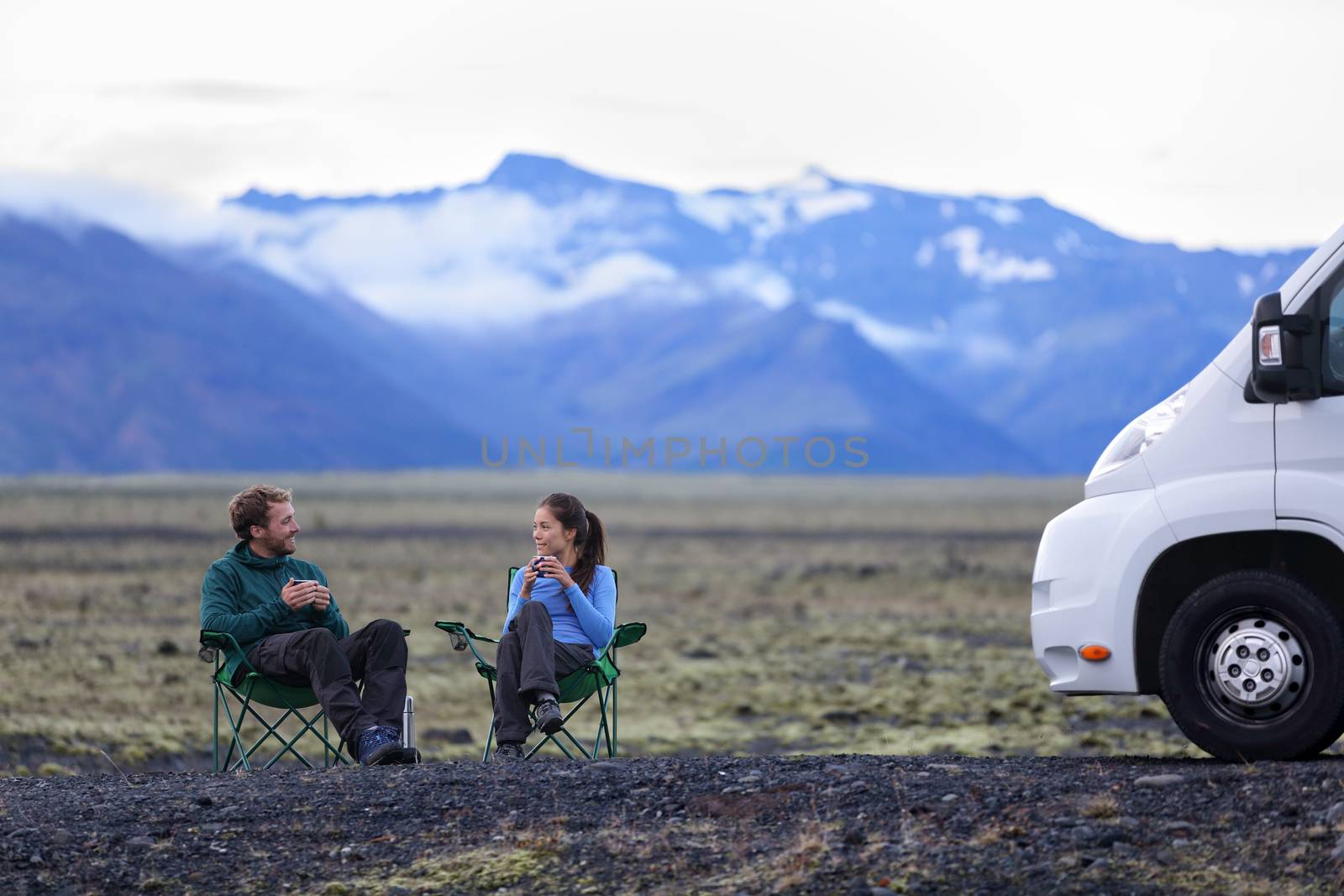 Travel couple by mobile motor home RV campervan. People sitting in chairs relaxing camping and enjoying traveling on Iceland in recreational vehicle. Young couple enjoying coffee in nature landscape.