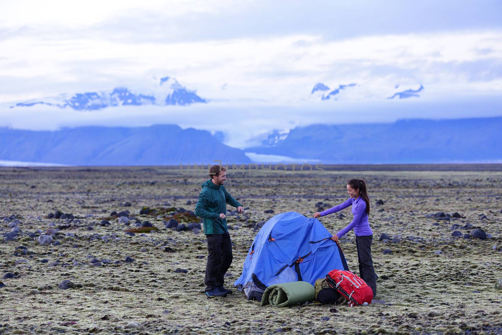 Camping couple pitching tent after hiking. Friends putting up tent for the night after hike with gear and backpacks. Active lifestyle image with Caucasian man and Asian woman.