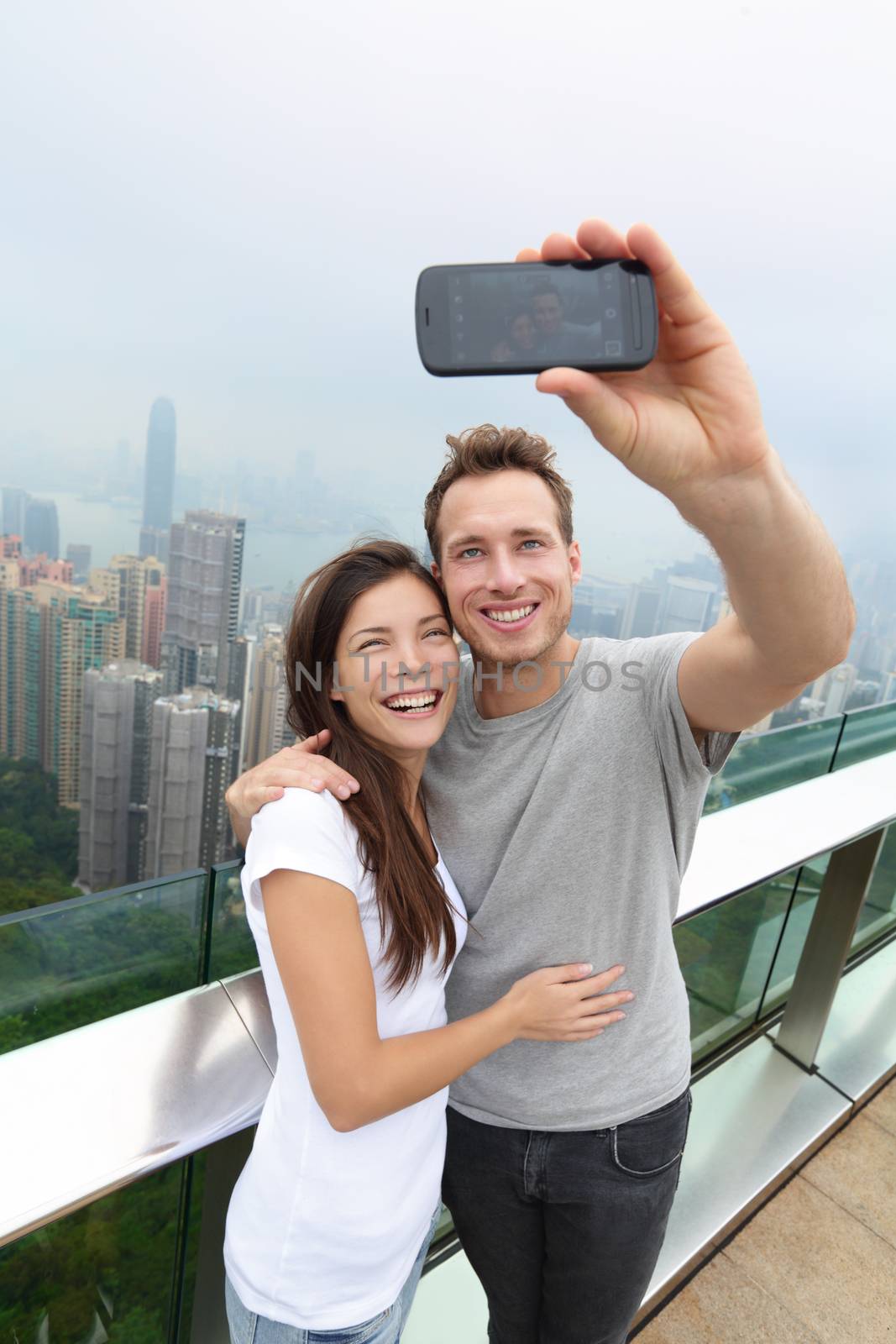 Hong Kong Victoria Peak tourists couple taking selfie photo picture with smartphone enjoying view over Hong Kong and Victoria Harbour. Young happy multiethnic couple traveling in Asia.