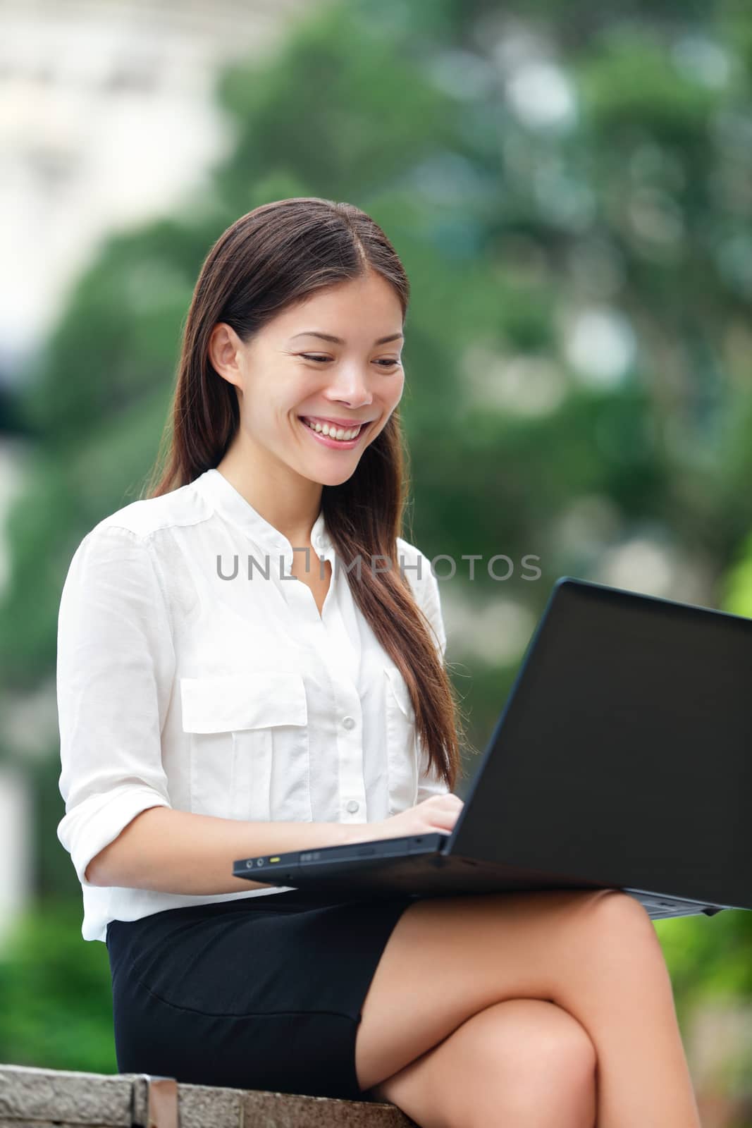 Businesswoman with computer laptop working outside looking at screen in business district, Central, Hong Kong. Young female professional business woman sitting outdoors. Asian Chinese Caucasian woman.