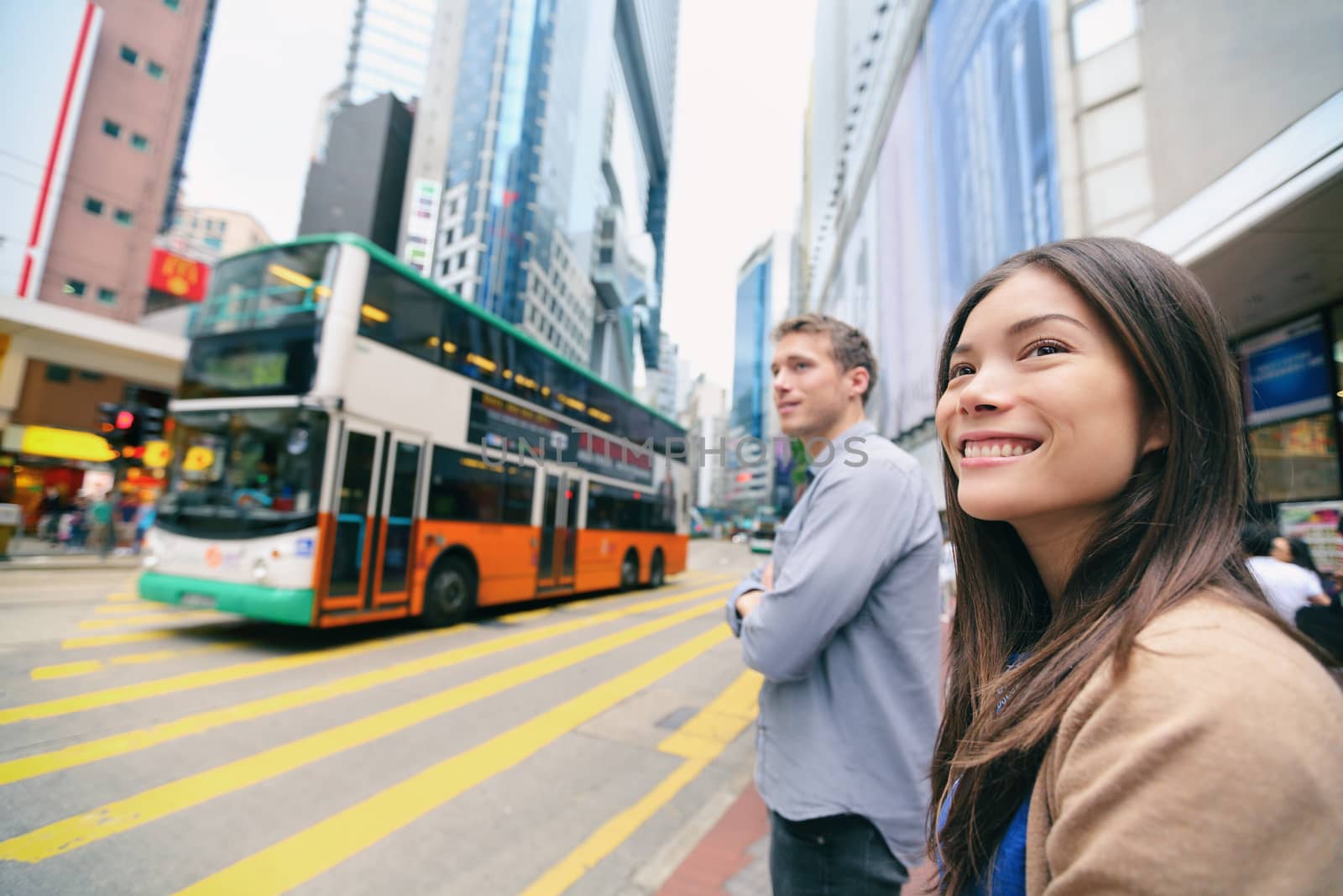 Hong Kong People walking waiting for traffic lights to cross busy road with double decker bus. Youn interracial Asian Chinese / Caucasian woman and man smiling happy living in city. by Maridav