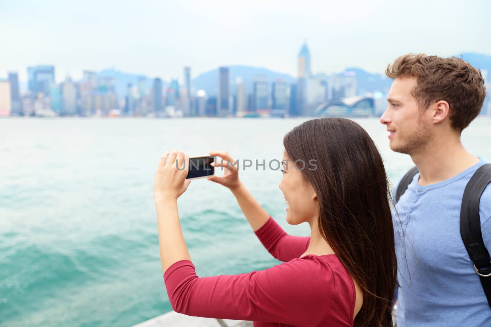 Tourist couple taking photo pictures enjoying view and sightseeing on Tsim Sha Tsui Promenade and Avenue of Stars in Victoria harbour, Kowloon, Hong Kong. Tourism travel concept.